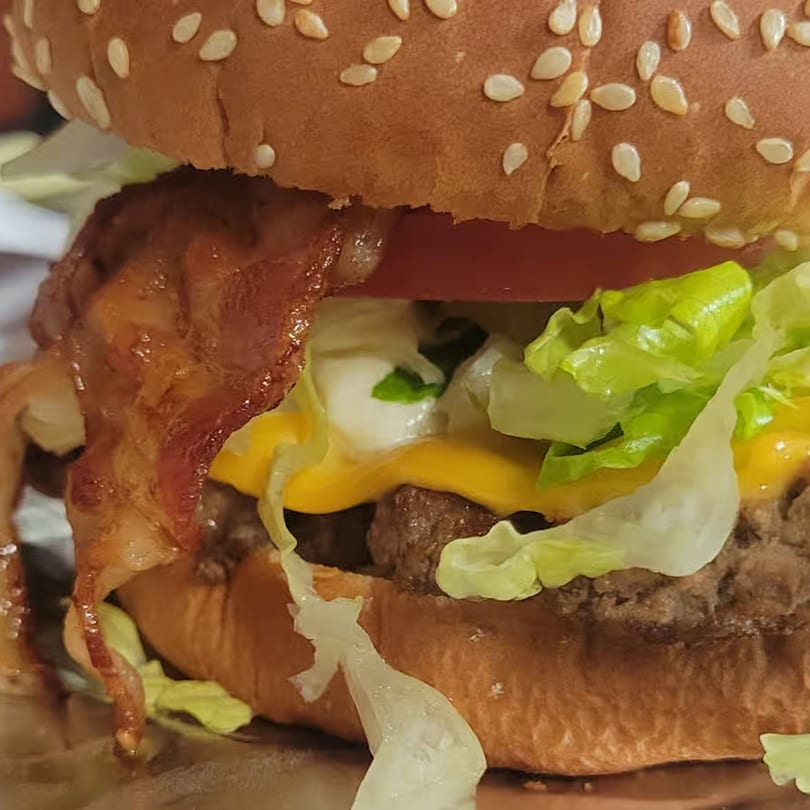 Close-up of a ChicknCoop burger with bacon, melted cheese, lettuce, and tomato on a sesame seed bun.